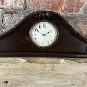Puede incluir: Un reloj de sobremesa de madera marrón oscuro con una silueta triangular arqueada. La esfera es circular con fondo blanco, números arábigos negros y dos manecillas negras. El marco de madera presenta detalles circulares tallados.