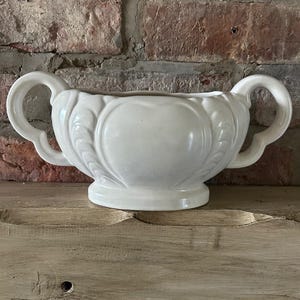 Pode incluir: Um vaso de cerâmica branco com duas alças curvas. O vaso tem um design decorativo em forma de coração no centro e assenta numa pequena base. O fundo apresenta uma parede de tijolo desgastada e uma superfície de madeira.