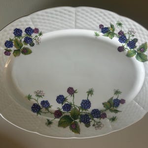 Puede incluir: Una fuente de servir ovalada de porcelana blanca con un borde texturizado de tejido de cesta y una ilustración botánica central de moras de color púrpura oscuro, hojas verdes y pequeñas flores blancas en tres grupos.
