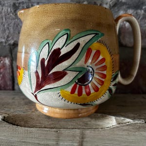May include: A ceramic pitcher with a tan and cream glazed finish featuring a prominent crackle texture. The body is decorated with hand-painted floral motifs in shades of deep red, yellow, and green. It includes a curved handle and a small spout.