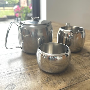 May include: A polished stainless steel tea set consisting of a teapot with a lid and handle, a creamer pitcher with a spout and handle, and an open sugar bowl. Each piece features a smooth, reflective metallic finish and a rounded, simple design.