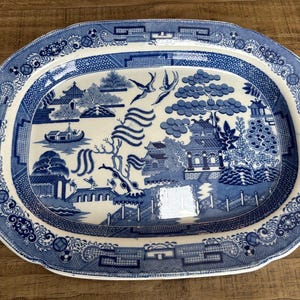 Pode incluir: Travessa oval de cerâmica azul e branca com um design detalhado do padrão Willow. A travessa apresenta uma cena central de paisagem com pagodes, pontes e pássaros, rodeada por uma borda decorativa. Uma peça clássica para servir.