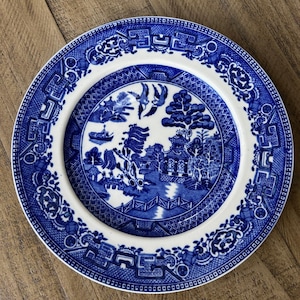 Puede incluir: Un plato redondo de cerámica azul y blanco con un diseño de patrón Willow. El plato presenta una escena central de un puente, edificios y pájaros, rodeada por un borde decorativo. El plato está sobre una superficie de madera.