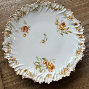 Może przedstawiać: Biały talerz porcelanowy z falistym, złoconym brzegiem. Powierzchnia ozdobiona jest delikatnym wzorem pomarańczowych i żółtych róż z zielonymi liśćmi, rozmieszczonych wokół środka oraz wzdłuż teksturowanego, żłobkowanego obrzeża talerza.