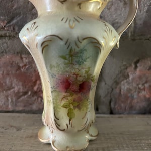 May include: A vintage, cream-coloured ceramic jug with a floral design. The jug features a flared base, a curved handle, and gold trim. The floral design includes pink and red flowers with green leaves.