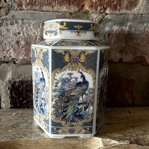 Op de afbeelding: Een zeshoekige porseleinen pot met een afneembaar deksel, voorzien van een donkerblauwe achtergrond met goudomrande panelen. Elk paneel toont een gedetailleerde illustratie van een pauw omringd door kleurrijke bloemen, waaronder roze en blauwe bloesems.