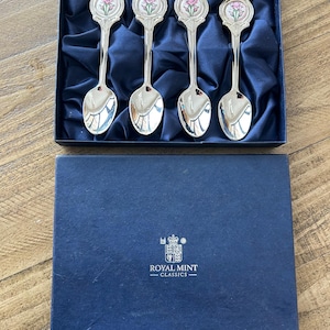 Può includere: Un set di quattro cucchiai d'argento con decorativi disegni di cardo in una scatola di presentazione blu. I cucchiai hanno un motivo floreale sul manico e sono inseriti in un tessuto simile al raso. La scatola ha il logo Royal Mint Classics.