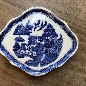 May include: A white and blue decorative plate with a scalloped edge. The plate features a blue and white Willow pattern design with a landscape scene, including buildings, trees, and birds. The plate is likely made of ceramic.