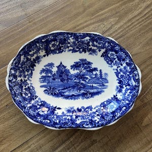 Może przedstawiać: Owalny ceramiczny półmisek z niebiesko-białym wzorem kwiatowym i krajobrazowym. Środek przedstawia szczegółową scenę z budynkami, drzewami i postaciami. Obrzeże ma falisty brzeg i jest ozdobione wzorami kwiatowymi.
