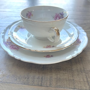 Könnte beinhalten: Ein weißes Porzellan-Teeservice, bestehend aus einer Tasse, einer Untertasse und einem Kuchenteller. Das Set hat ein zartes Blumenmuster in Rosa und Lila mit goldenen Verzierungen. Die Tasse hat einen geschwungenen Henkel.