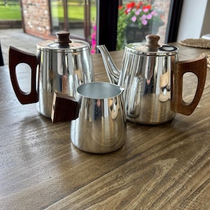 Puede incluir: Un juego de té de tres piezas que consta de una tetera, una cafetera y una jarra de leche. Cada pieza está fabricada en metal plateado pulido y cuenta con mangos de madera de color marrón oscuro y pomos a juego en las tapas de las ollas.
