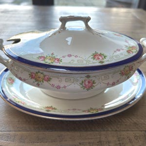 May include: A white porcelain soup tureen with a lid, handles, and a matching saucer. The tureen and saucer are decorated with a blue border and floral patterns in pink, yellow, and green. The tureen has a handle on the lid.