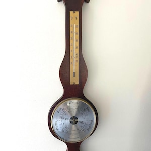 May include: A vintage wooden weather station with a thermometer, barometer, and hygrometer. The thermometer displays both Celsius and Fahrenheit, while the barometer indicates changing weather conditions. The wooden frame is a dark brown color.