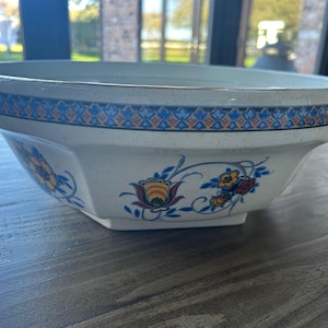 Könnte beinhalten: Eine cremefarbene, achteckige Keramikschale mit einem dekorativen blau-orangen Blumenmuster. Der Rand hat ein blau-oranges geometrisches Design. Die Schale steht auf einer Holzoberfläche.