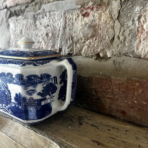 Peut inclure: Un sucrier en porcelaine blanche orn&eacute; d'un motif bleu de type willow repr&eacute;sentant des paysages traditionnels, avec un couvercle carr&eacute; surmont&eacute; d'un petit bouton et des bords dor&eacute;s. Le sucrier poss&egrave;de une anse lat&eacute;rale et une finition brillante.