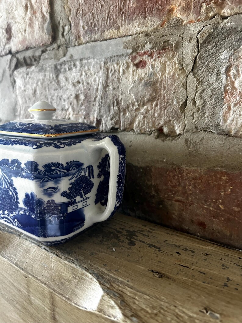 Peut inclure: Un sucrier en porcelaine blanche orn&eacute; d'un motif bleu de type willow repr&eacute;sentant des paysages traditionnels, avec un couvercle carr&eacute; surmont&eacute; d'un petit bouton et des bords dor&eacute;s. Le sucrier poss&egrave;de une anse lat&eacute;rale et une finition brillante.