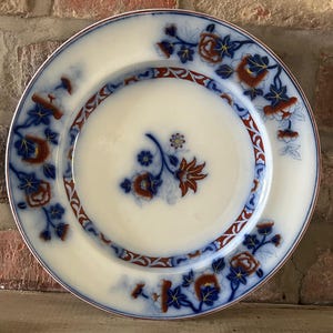 Op de afbeelding: Witte keramische plaat met een bloemmotief in blauw, rood en goud. De plaat heeft een centraal bloemmotief en een decoratieve rand. De rand is zachtroze. Een decoratieve plaat in vintage stijl.