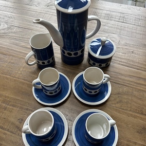 以下が含まれることがあります： 青と白のセラミックコーヒーセット。セットには、背の高いコーヒーポット、クリーマー、砂糖入れ、ソーサー付きのカップが4つ含まれています。デザインは、幾何学模様と白いアクセントが付いた青いバンドが特徴です。木製の表面に配置されています。