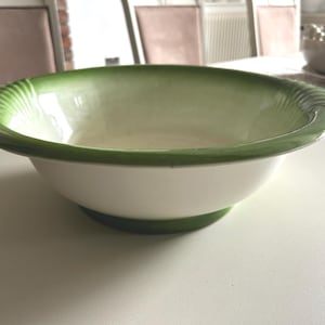 May include: A white ceramic bowl with a green rim and base. The rim has a decorative, ridged design. The interior of the bowl is a light green color. The bowl is sitting on a white surface.