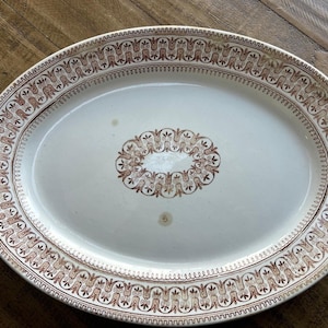 May include: Oval serving platter with a cream-colored surface and a brown decorative border. The border features a repeating pattern of stylized floral and scroll elements. A similar design is in the center of the platter.