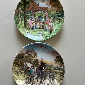 May include: Two decorative plates featuring detailed rural scenes. One plate shows a gathering outside a house, with people and geese. The other plate depicts a man riding a horse, with other people and dogs. The plates have green and blue borders.