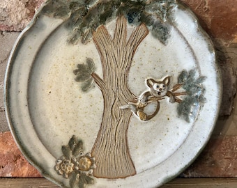May include: A ceramic plate with a tree and koala design. The plate has a cream-colored center with a textured brown tree trunk and a koala clinging to a branch. The rim is a mottled green, and there are floral accents.