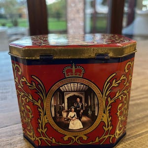 May include: Vintage red and gold tin with a royal portrait. The tin has a red background with gold filigree and a central image of a royal couple. The lid and edges are gold and blue.