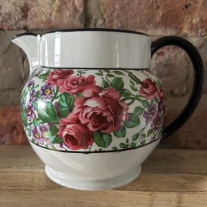 May include: A white ceramic pitcher featuring a floral pattern of red roses and small purple flowers around the center. The pitcher has a black handle, a black rim, and a black horizontal line separating the floral band from the white base.