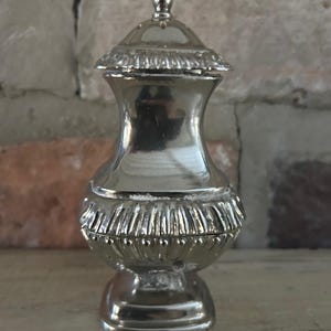 Puede incluir: Un pequeño tarro decorativo plateado con tapa. El tarro tiene un cuerpo bulboso con un diseño detallado y texturizado alrededor de la base y la tapa. La tapa está rematada con un pequeño remate redondo. El tarro se exhibe sobre una superficie de madera.