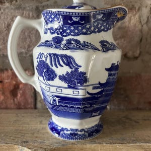 May include: A white porcelain creamer with a blue willow pattern featuring pagodas, trees, and a bridge. The creamer has a matching lid with a gold-trimmed finial, a curved handle, and a spout. The surface shows fine crackle glazing throughout.