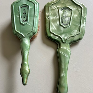 May include: Two vintage green vanity brushes. The brushes have a marbled effect and a shield-shaped top with a black emblem. One brush has bristles. The handles are long and slender, with a pointed end. The brushes are displayed on a neutral background.