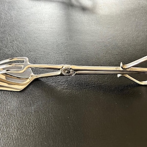 Puede incluir: Pinzas de servir plateadas con cabezal en forma de pala y mango con extremos en bucle. Un diseño floral decora el punto de giro. Las pinzas son probablemente para pasteles o repostería. Miden unos 20 cm de largo.