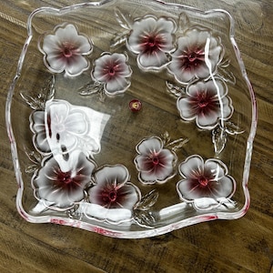 May include: A square clear glass serving tray with scalloped edges, featuring a decorative pattern of frosted white flowers with deep red centers. The glass surface is smooth and transparent, highlighting the floral motifs and a small red circular emblem.
