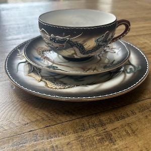 May include: A porcelain tea cup and saucer set featuring a black and white gradient background with a detailed dragon illustration. The cup has a dark handle with gold trim, and both pieces display a raised dragon motif with blue and gold accents.