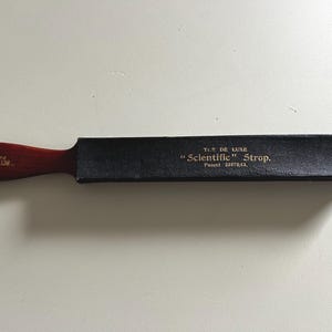 Op de afbeelding: Een vintage leren riem met een donkerhouten handvat. Het handvat is ovaal van vorm en heeft de woorden "SCIENTIFIC PATENT" erop gedrukt. De zwarte leren riem heeft de woorden "T.S. DE LUXE" en "Scientific Strop" erop gedrukt.