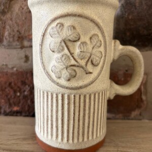 May include: A cream-coloured ceramic mug with a brown base and handle. The mug features a circular embossed design of a shamrock and vertical lines. The mug is approximately 13 cm tall.