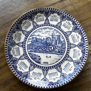 May include: A round white ceramic plate with a blue transferware pattern featuring Windsor Castle in the center, the year 1984, and a calendar for each month arranged around the border. The rim is decorated with blue floral motifs and gold trim.