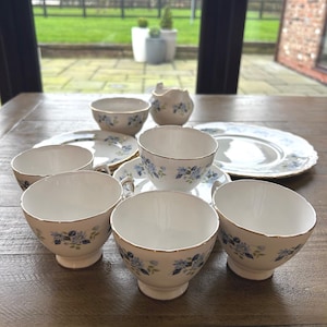 May include: A floral patterned tea set. The set includes six teacups, saucers, a sugar bowl, and a serving plate. The teacups and saucers have a white base with blue floral designs and gold trim. The set is arranged on a wooden table.