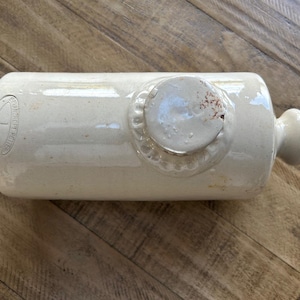 Peut inclure: Bouteille en grès cylindrique de couleur crème, avec une extrémité arrondie et une petite poignée. La bouteille présente un motif circulaire en relief sur le dessus. Le texte "GLASGOW" et "SCOTTISH POTTERS" est visible sur le côté.