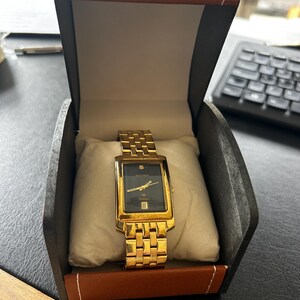Op de afbeelding: Een rechthoekig goudkleurig polshorloge met een zwarte wijzerplaat, gouden wijzers en een datumvenster, gepresenteerd op een wit kussen in een bruine en zwarte doos. De wijzerplaat bevat de tekst Sekonda en een kleine diamant op twaalf uur.
