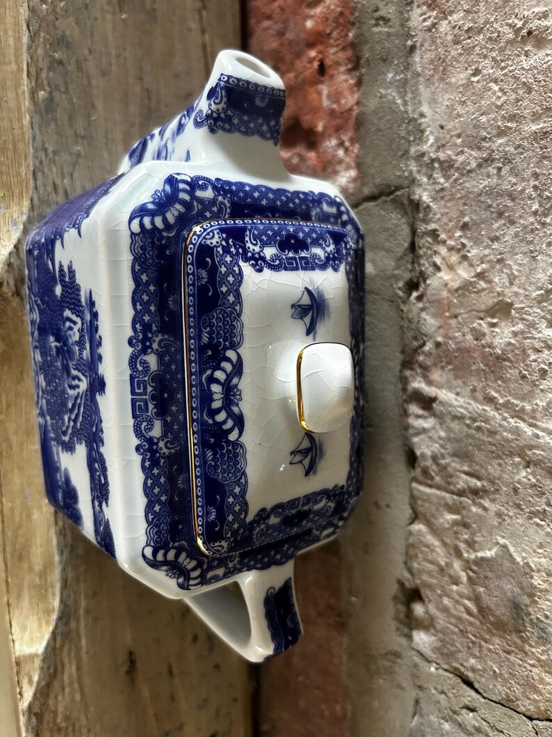 Peut inclure: Une th&eacute;i&egrave;re rectangulaire en c&eacute;ramique avec une base blanche orn&eacute;e de motifs bleus complexes de style willow et une finition &eacute;maill&eacute;e craquel&eacute;e. Le couvercle poss&egrave;de un petit bouton blanc avec un bord dor&eacute;, assorti &agrave; la bordure dor&eacute;e du couvercle.