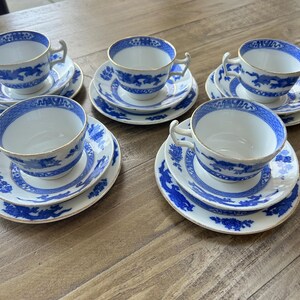 Puede incluir: Un juego de cinco tazas de té de porcelana blanca con sus platos a juego, decoradas con un patrón de dragón azul y borde dorado. Las tazas presentan una forma de asa distintiva y los platos muestran un motivo floral y de dragón azul.