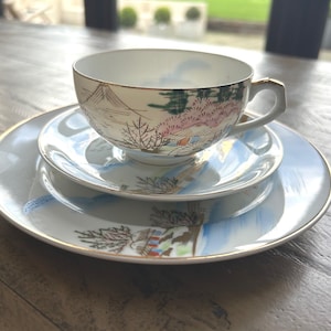 May include: A vintage teacup, saucer, and side plate set. The porcelain set features a landscape design with mountains, trees, and buildings in green, pink, and blue. The edges are trimmed with gold.