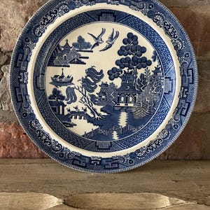 May include: A blue and white ceramic plate with a Willow pattern design. The plate has a central landscape scene with birds, trees, and buildings, and a decorative border. The plate is set against a rustic brick background, typical of a traditional kitchen.