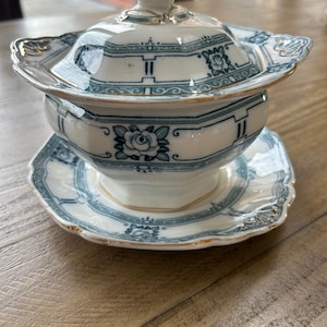 Puede incluir: Una sopera de porcelana blanca y verde azulado con tapa y platillo a juego. La sopera presenta un diseño floral y geométrico con ribetes dorados. La tapa tiene un adorno decorativo. El juego está colocado sobre una superficie de madera.
