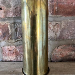 May include: A tall, cylindrical brass vase with a slightly flared top and a circular base. The vase has a polished, reflective surface, showing hints of wear. The background features a rustic brick wall and wooden surface.