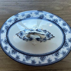 Op de afbeelding: Ovale serveerschaal met deksel. De witte porseleinen schaal heeft een blauw bloemmotief en een decoratieve handgreep. De rand is versierd met een Grieks sleutelontwerp. De schaal is ongeveer 30 cm lang.