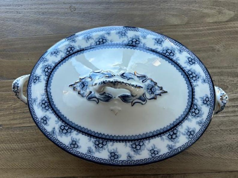 Op de afbeelding: Ovale serveerschaal met deksel. De witte porseleinen schaal heeft een blauw bloemmotief en een decoratieve handgreep. De rand is versierd met een Grieks sleutelontwerp. De schaal is ongeveer 30 cm lang.
