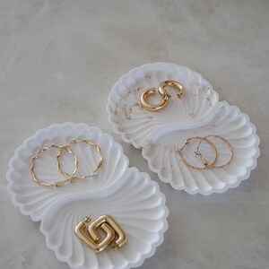 Op de afbeelding: Twee witte, schelpvormige trays met gouden sieraden. Eén tray bevat twee paar gouden oorringen en de andere een paar gouden vierkante oorbellen. De trays hebben een marmerachtig patroon.