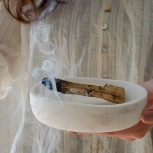 Könnte beinhalten: Eine weiße Keramikschale enthält ein glühendes Stück Palo Santo-Holz, das Rauchschwaden abgibt. Die Schale wird vor einer Person gehalten, die ein weißes Hemd mit Knöpfen trägt. Die Szene vermittelt ein Gefühl von Ruhe und spiritueller Praxis.